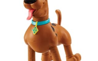 A plastic Scooby-Doo toy figure stands with a happy expression, tongue out and eyes wide. It has brown fur, a blue collar with a yellow diamond-shaped tag, and black spots on its back. The figure’s legs are slightly bent as if in mid-step.