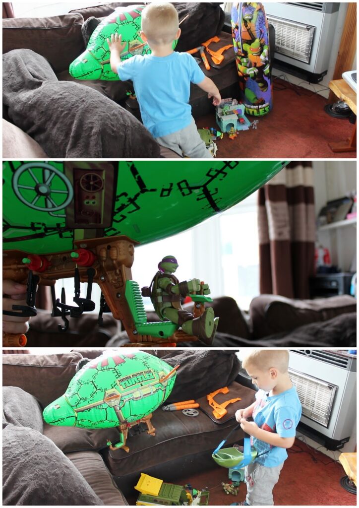 A young boy with short blond hair wearing a light blue shirt plays with a large green Teenage Mutant Ninja Turtles blimp toy on a brown sofa. Various TMNT action figures and accessories are scattered around him in a brightly lit lounge.