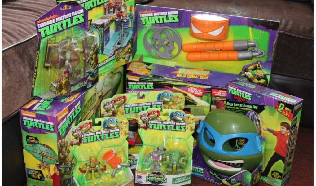 A collection of Teenage Mutant Ninja Turtles toys and action figures in colourful packaging is arranged on a brown sofa. The toys include weapons, action figures, and a blue TMNT mask, all displaying the TMNT logo and vibrant cartoon artwork.