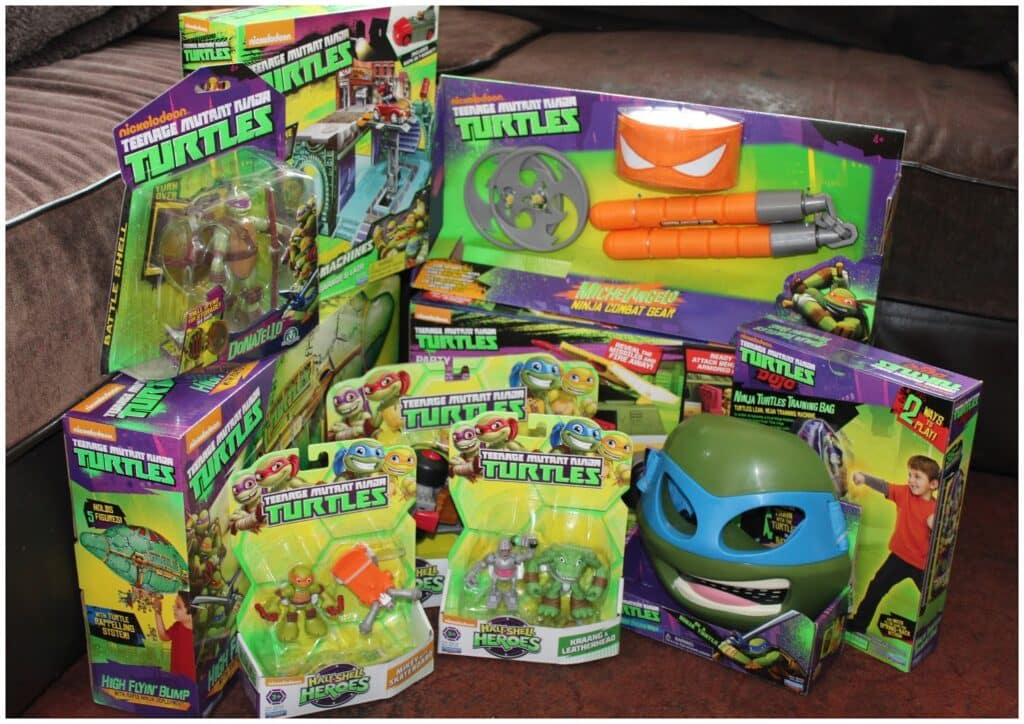 A collection of Teenage Mutant Ninja Turtles toys is displayed on a sofa. The items include action figures, nunchaku, a mask, and other themed accessories, all in colourful, predominantly green and purple packaging featuring cartoon turtle graphics.