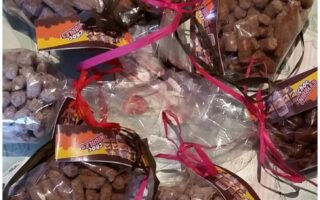 Assorted clear bags filled with brown, stick-shaped snacks, tied with ribbons and colourful tags, arranged in a circular pattern on a decorative tablecloth with cupcake illustrations and handwritten-style text.