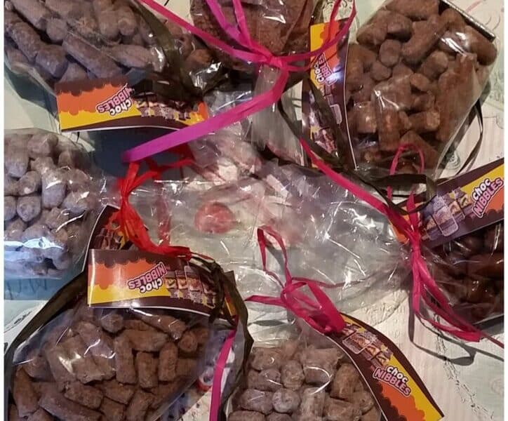 Assorted clear bags filled with brown, stick-shaped snacks, tied with ribbons and colourful tags, arranged in a circular pattern on a decorative tablecloth with cupcake illustrations and handwritten-style text.