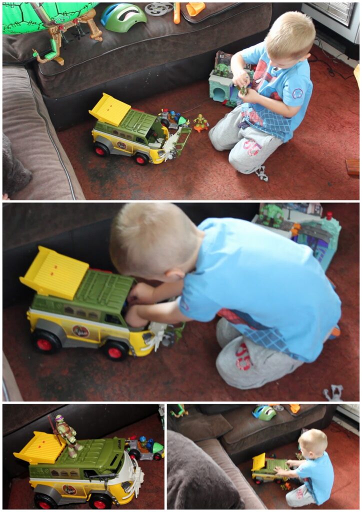 A young boy with blond hair plays on a carpeted floor with a large yellow toy lorry and several action figures, including a Teenage Mutant Ninja Turtle. He kneels and interacts with the toys, surrounded by other small toys and furniture.