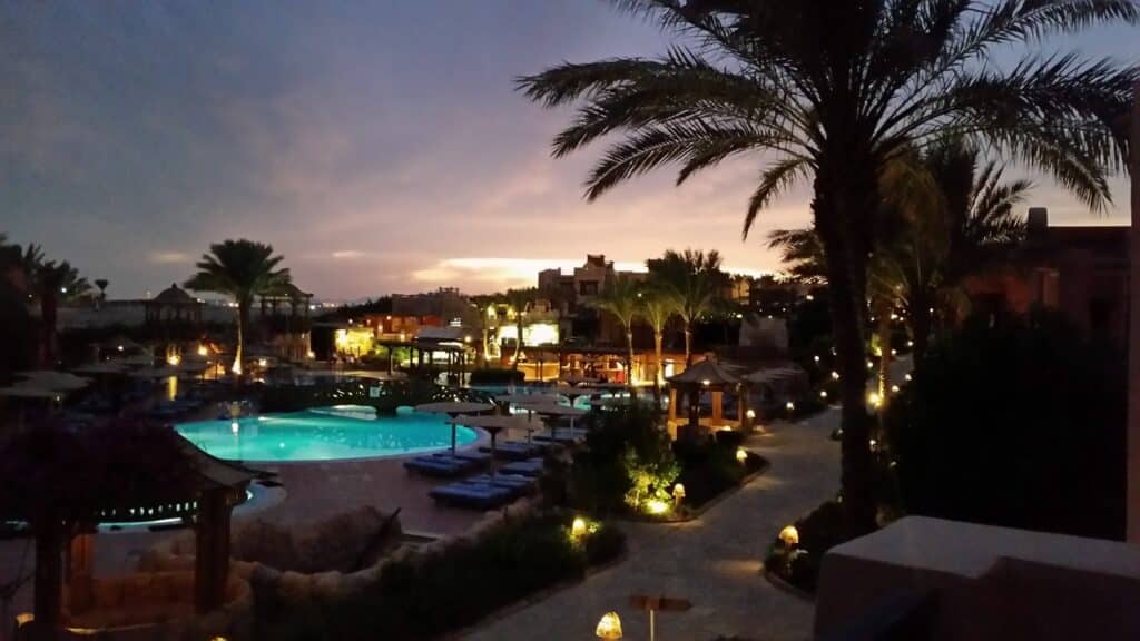 A resort at dusk with illuminated pools and pathways, sun loungers and parasols around the pool, palm trees lining the area, and buildings in the background. The sky is dusky purple and orange, creating a serene, tropical atmosphere.
