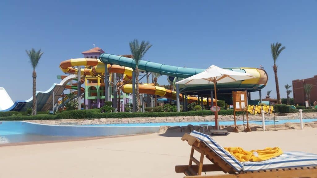 A waterpark scene with tall, colourful waterslides, palm trees, and a lazy river. In the foreground, a wooden lounger with blue-striped cushions and a white umbrella sits on sandy ground under a clear blue sky.