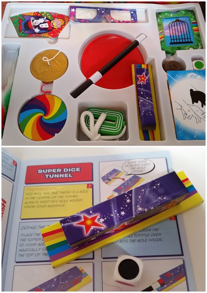 A children's magic kit with colourful props—magic wand, rope, giant coin, rainbow spinner, red disc, green frames, and decorated boxes—arranged in a tray. Below, one prop rests on a comic-style instruction booklet titled "SUPER DICE TUNNEL.
