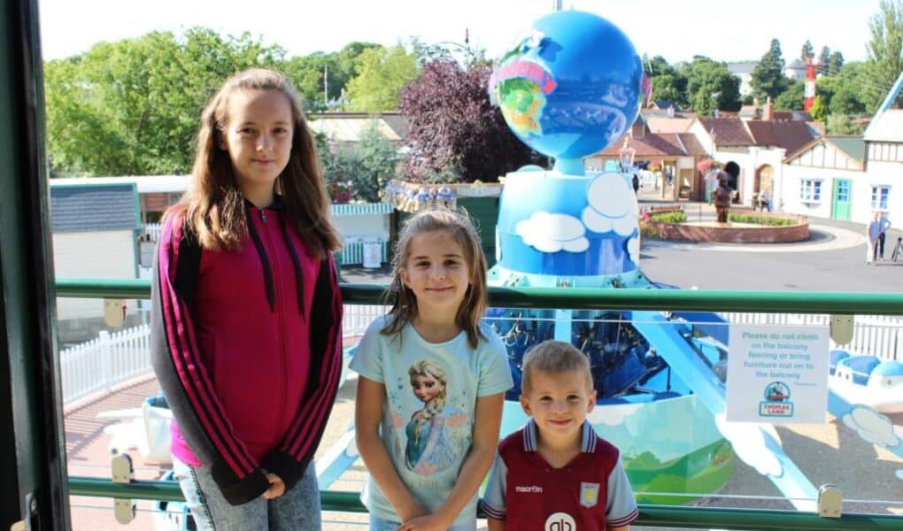 Three children stand smiling in front of a colourful amusement park ride with blue and white globes. Behind them is a white fence, greenery, and small buildings under a sunny sky. A sign on the railing asks visitors not to climb or lean.