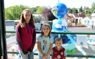 Three children stand smiling in front of a colourful amusement park ride with blue and white globes. Behind them is a white fence, greenery, and small buildings under a sunny sky. A sign on the railing asks visitors not to climb or lean.