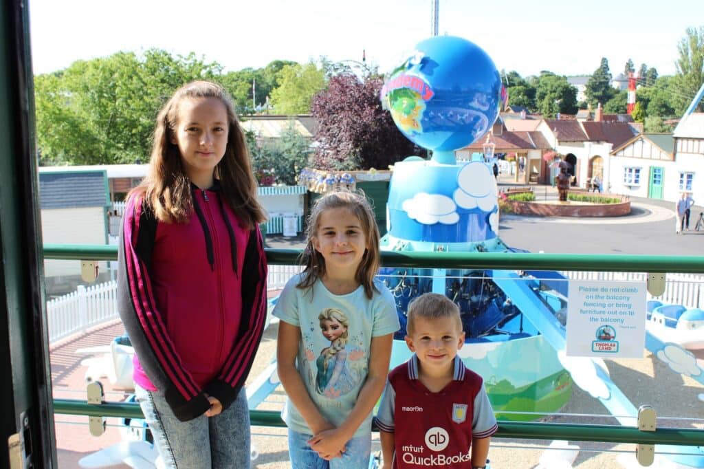 Three children stand smiling in front of a railing at an amusement park, with a blue spinning globe ride behind them. Two girls (one in a pink jacket, one in an Elsa top) and a boy in a maroon football shirt pose together on a sunny day.