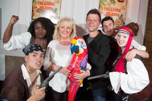 Five adults pose indoors, four dressed as pirates with bandanas, striped headbands, and fake weapons. A blonde woman in pink stands in the centre, holding a colourful inflatable parrot. Two "Treasure Island" posters are on the wall behind them.