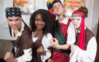 Four young adults in pirate costumes pose fiercely, flexing their arms. They wear bandanas, sashes, and shirts typical of pirates. A colourful parrot prop is perched behind them. Posters on the wall read “Treasure Island,” suggesting they are at a themed event or play.