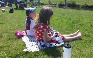 Two children sit barefoot on a blanket in the grass, eating snacks. The girl wears a red polka-dot dress with a Minnie Mouse print, and the boy wears a cap and sports shirt. A water bottle is in the foreground; people and a stream are in the background.