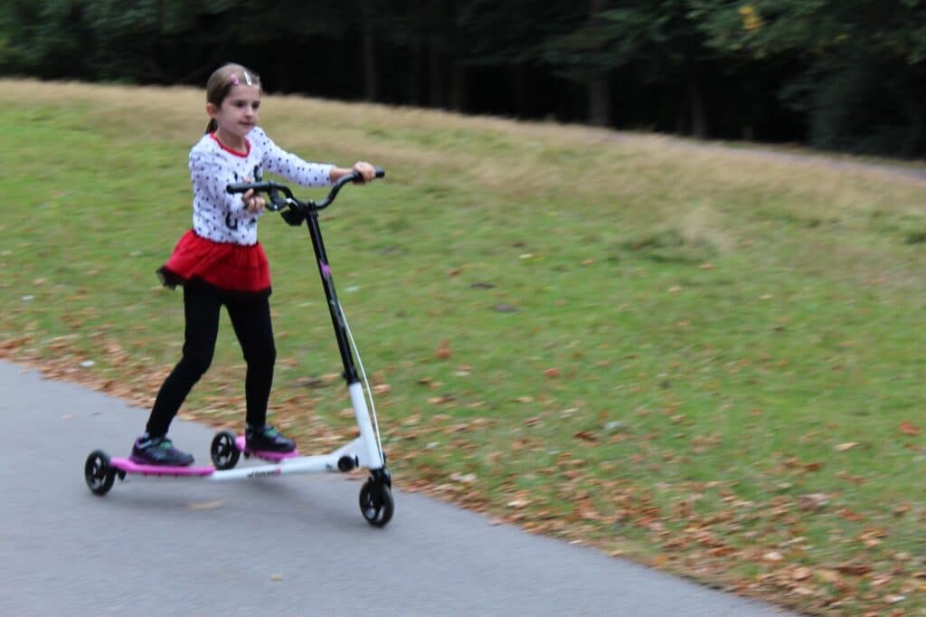A young girl wearing a red skirt, white jumper with black patterns, and black leggings rides a three-wheeled scooter on a paved path in a park. Green grass and some brown leaves are visible, with trees in the background.