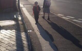 Two children carrying rucksacks walk on a sunlit street near the kerb, casting long shadows. Houses line the road, several cars are parked, and the bright sunlight creates a haze, making details in the background less distinct.