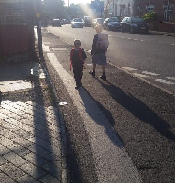 Two children carrying rucksacks walk on a sunlit street near the kerb, casting long shadows. Houses line the road, several cars are parked, and the bright sunlight creates a haze, making details in the background less distinct.