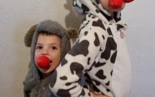 Two young children pose back-to-back. One wears a furry grey costume with a hood and red clown nose, the other wears a cow-print onesie with attached hood and two red clown noses in their mouth, facing sideways with a hand on their hip. A white textured wall is behind them.