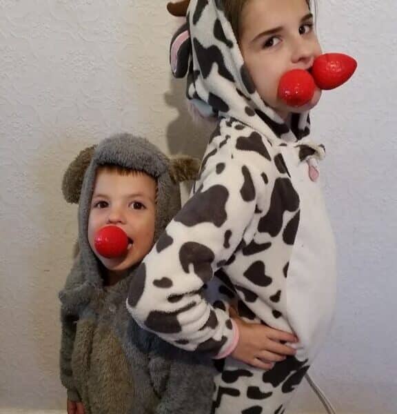 Two young children pose back-to-back. One wears a furry grey costume with a hood and red clown nose, the other wears a cow-print onesie with attached hood and two red clown noses in their mouth, facing sideways with a hand on their hip. A white textured wall is behind them.