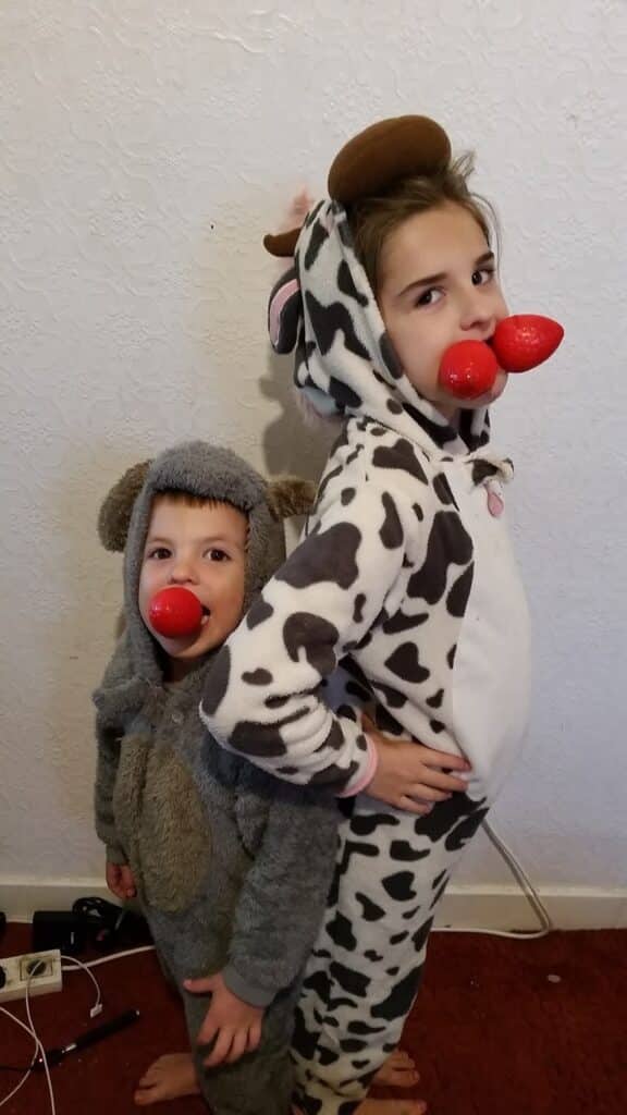 Two children stand back to back. One wears a cow onesie; the other wears a grey animal onesie. Both have red clown noses in their mouths and playful expressions. The background is a white textured wall, with some electrical cords and plugs visible near their feet.