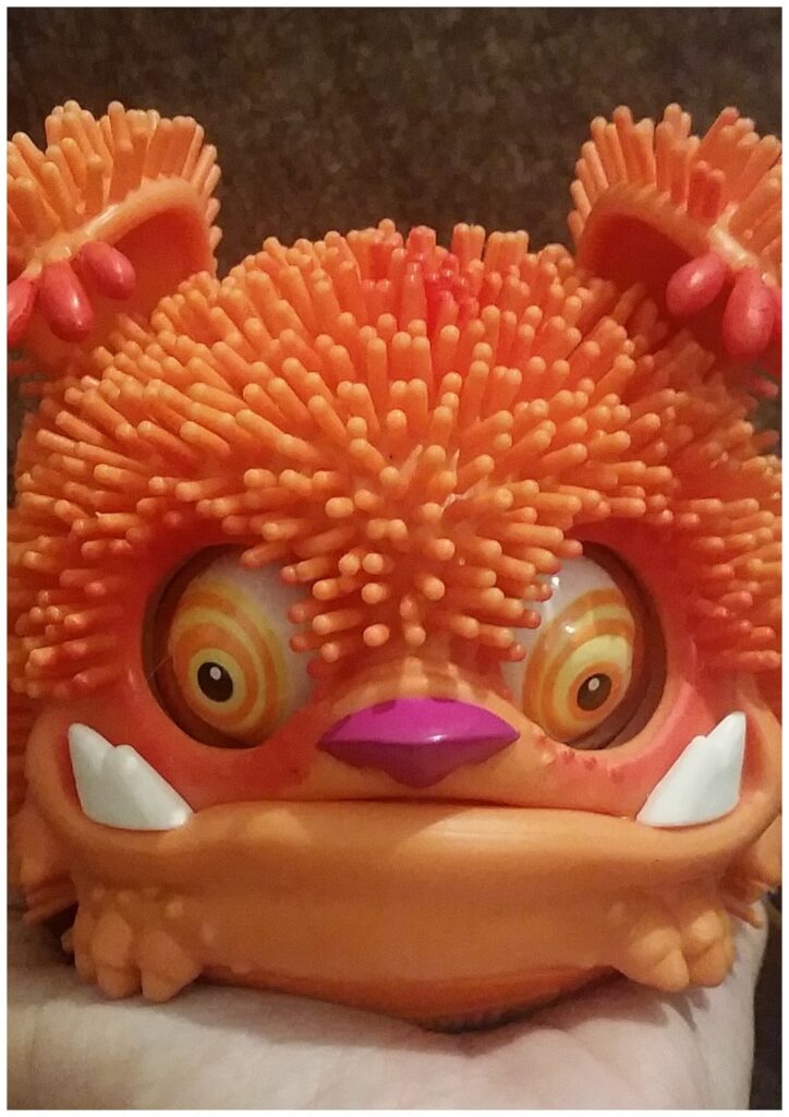 A close-up of an orange, fuzzy, monster toy with textured spikes, large swirling eyes, exaggerated white fangs, and a purple nose. The toy has bear-like ears and a slightly grumpy or surprised expression, held in a person's hand against a neutral background.