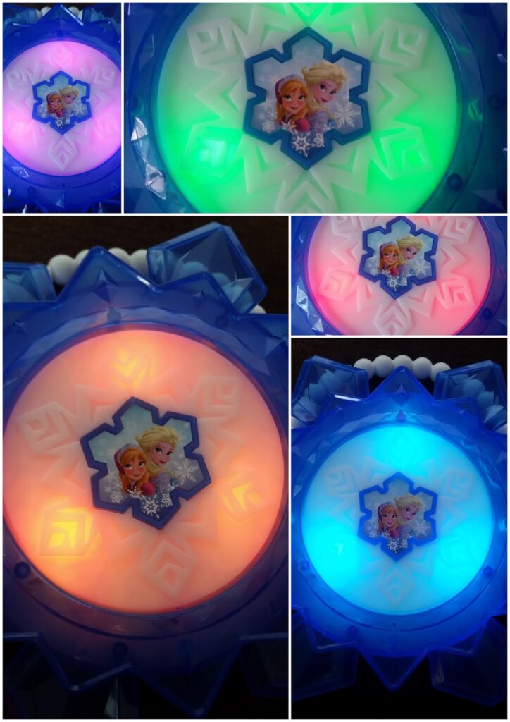 A blue, snowflake-shaped Frozen-themed toy features images of Elsa and Anna in the centre. The toy lights up in various colours—green, pink, orange, and blue—shown in five close-up photos with the sisters framed by a white snowflake pattern.