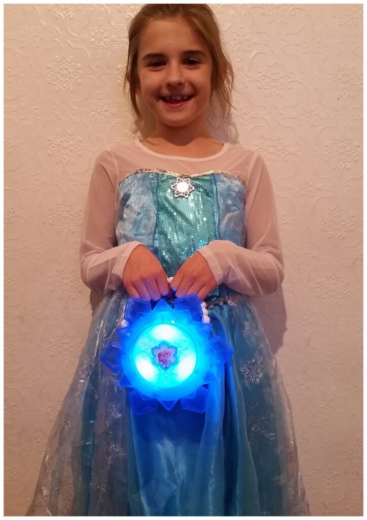 A young girl smiles at the camera, dressed in a sparkly blue and white Elsa costume from Frozen. She holds a glowing blue round handbag with a snowflake design and Elsa's image on it. The background is a textured white wall.