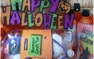 A colourful, glittery “Happy Halloween” sign with a pumpkin decorates a basket filled with orange bottles, a “Bumper Craft Tub” from Hobbycraft, and a Halloween card, all arranged with festive, spooky decorations for Hallowe'en.