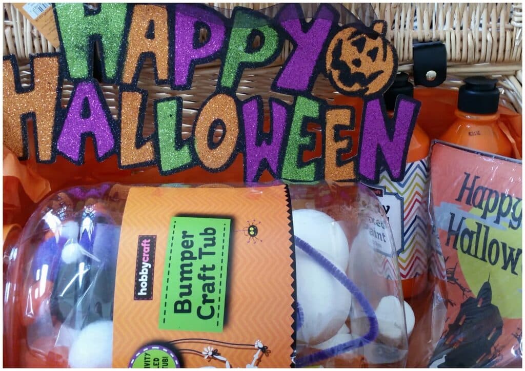 A colourful “Happy Halloween” sign with glittery letters and a pumpkin is displayed above a wicker basket. In front, there’s a Hobbycraft Bumper Craft Tub, orange bottles, and a card with chevron stripes and “Happy Hallow” text in purple and orange.