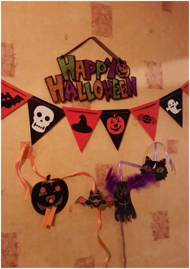 A wall decorated for Hallowe’en with a colourful "Happy Hallowe’en" sign, a triangular banner featuring a skull, witch’s hat, jack-o’-lantern, bat, and ghost, plus paper Hallowe’en crafts below: a pumpkin, bat, and purple-winged bat on orange ribbons.