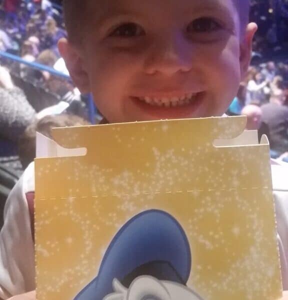 A young boy smiles brightly, holding a yellow container with a large image of Donald Duck’s face on it. He sits in an indoor arena filled with a crowd of people seated in the background, under bright blue and purple lights.