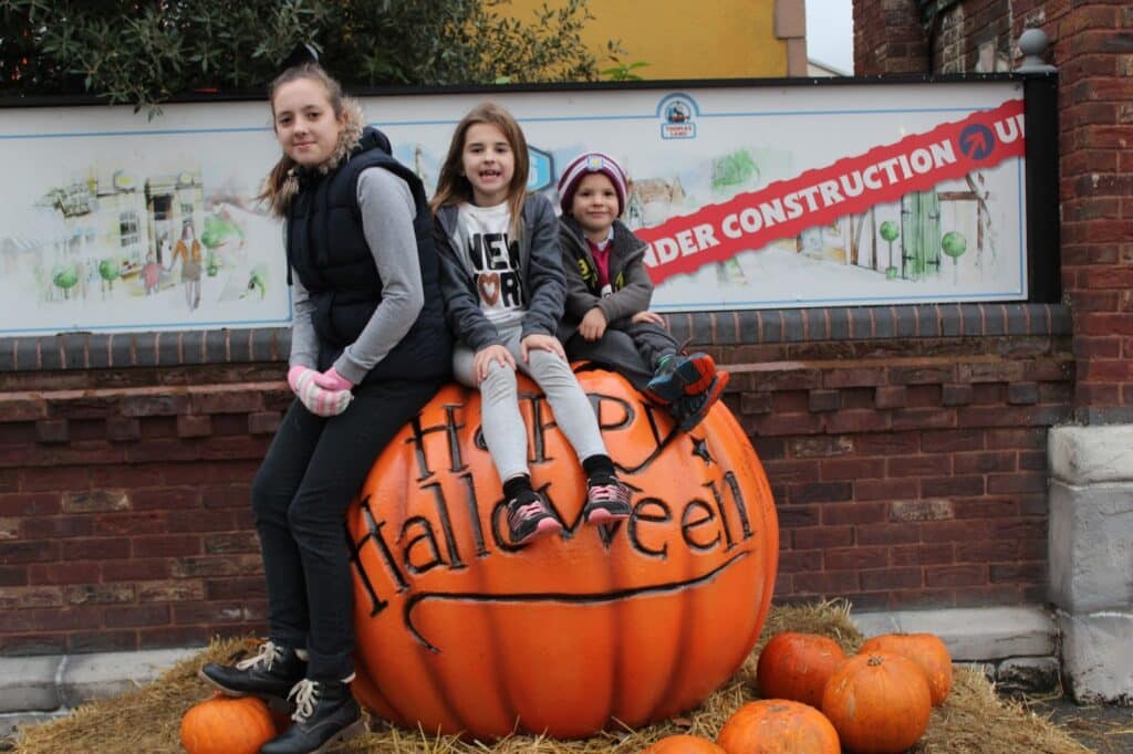 Three children sit on a large orange pumpkin with “Happy Halloween” written on it. They are bundled in jackets and gloves. Smaller pumpkins and straw surround the base. A brick wall and an “Under Construction” sign are in the background.