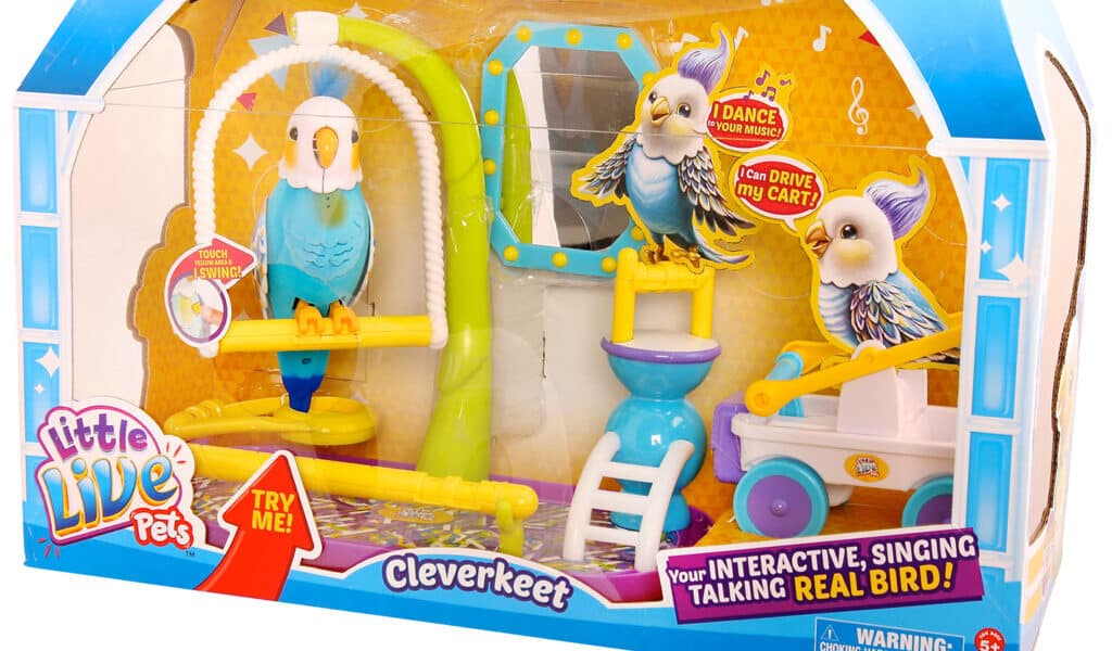 A “Little Live Pets Cleverkeet” toy set in packaging, showing a blue and yellow interactive toy bird on a perch, a swing, mirror, food dish, and a yellow trolley. The packaging features bright colours, logos, and text promoting the bird’s singing and talking abilities.
