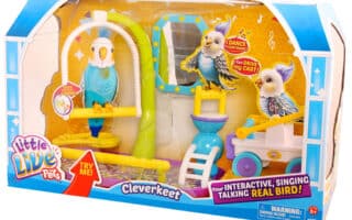 A “Little Live Pets Cleverkeet” toy set in packaging, showing a blue and yellow interactive toy bird on a perch, a swing, mirror, food dish, and a yellow trolley. The packaging features bright colours, logos, and text promoting the bird’s singing and talking abilities.
