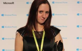 A woman with long brown hair wearing a black dress, green lanyard, and bracelets holds a glass of water. She stands in front of a step-and-repeat backdrop featuring the Windows Phone and Microsoft logos. The setting appears to be a tech event or party.