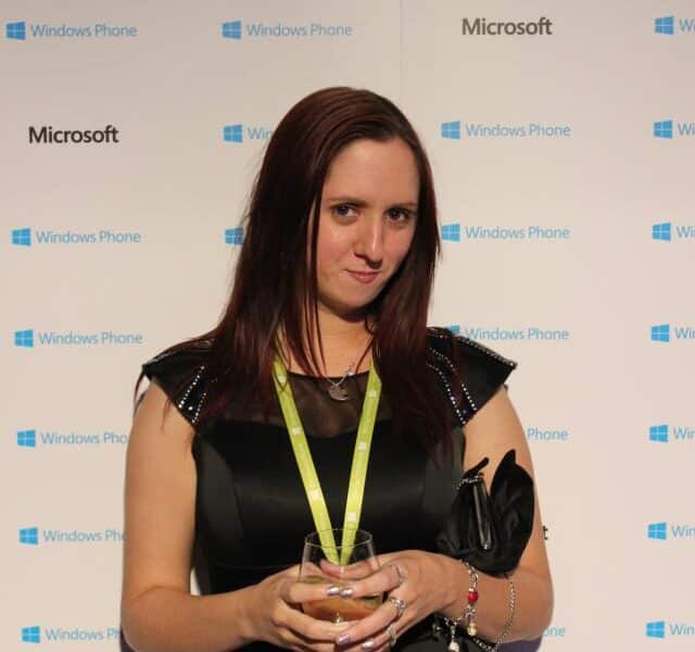 A woman with long brown hair wearing a black dress, green lanyard, and bracelets holds a glass of water. She stands in front of a step-and-repeat backdrop featuring the Windows Phone and Microsoft logos. The setting appears to be a tech event or party.