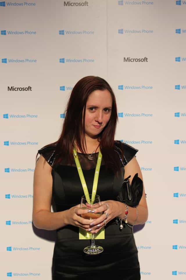 A woman with long brown hair wearing a black dress, green lanyard, and bracelets holds a glass of water. She stands in front of a step-and-repeat backdrop featuring the Windows Phone and Microsoft logos. The setting appears to be a tech event or party.