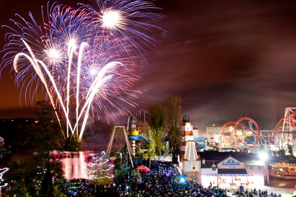 Colourful fireworks burst in the night sky above a brightly lit amusement park with roller coasters and buildings. A large crowd gathers below, watching the display. The scene is lively, with vibrant lights reflecting off the water and festive energy throughout.