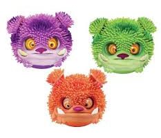 Three round, fuzzy-textured toy monster heads are shown: one purple with yellow eyes and small ears, one green with orange and yellow eyes, and one orange with red ears and yellow eyes. Each has a toothy grin and spiky, rubbery hair.