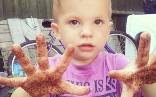 A young boy with short blond hair holds out his hands, covered in dirt, towards the camera. He wears a purple shirt with black text and stands outdoors near a bicycle, a fence, a tyre, and other items in the background.