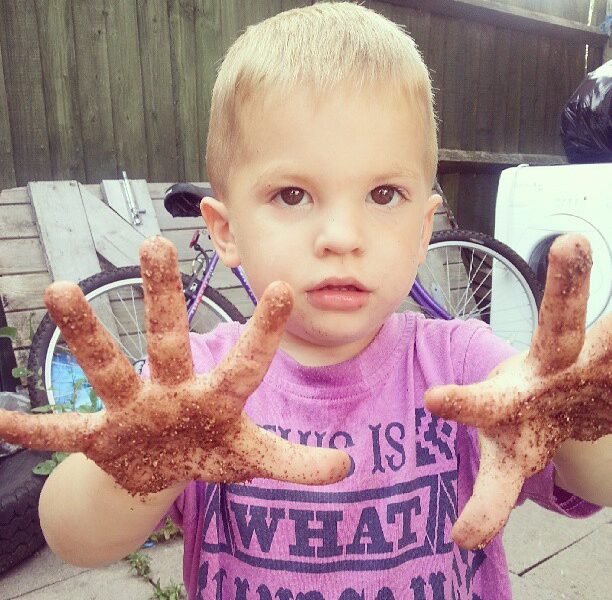A young boy with short blond hair holds out his hands, covered in dirt, towards the camera. He wears a purple shirt with black text and stands outdoors near a bicycle, a fence, a tyre, and other items in the background.