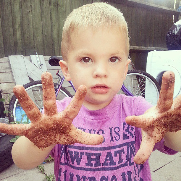 A young boy with short blond hair holds out his hands, covered in dirt, towards the camera. He wears a purple shirt with black text and stands outdoors near a bicycle, a fence, a tyre, and other items in the background.