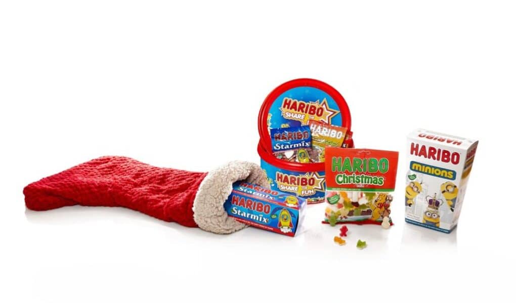A red Christmas stocking lies on its side, with assorted Haribo sweet packs spilling out, including Haribo Starmix, Haribo Christmas, and Haribo Minions, all set against a clean white background. Some gummy sweets are scattered near the packs.
