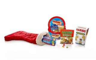 A red Christmas stocking lies on its side, with assorted Haribo sweet packs spilling out, including Haribo Starmix, Haribo Christmas, and Haribo Minions, all set against a clean white background. Some gummy sweets are scattered near the packs.