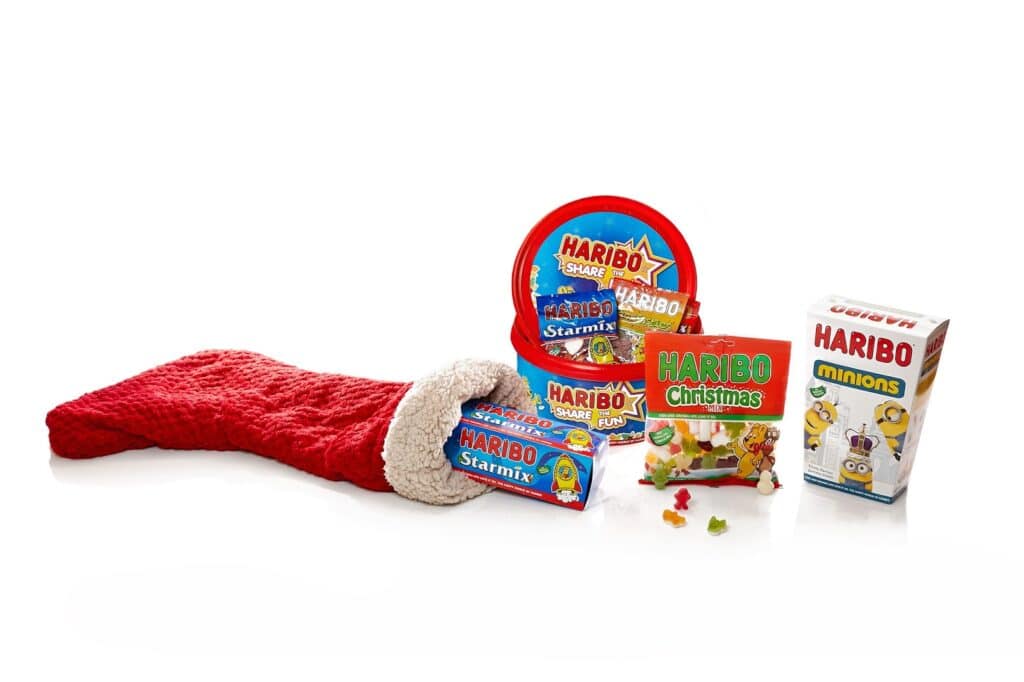 A red Christmas stocking lies on its side, spilling various Haribo sweet packages: Starmix in a round tub, Haribo Christmas gummies, Haribo Minions, and another Starmix pack. Several colourful gummy sweets are scattered in front.