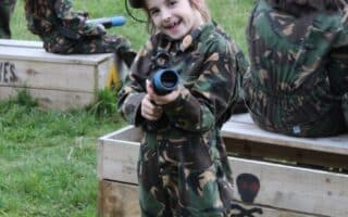 A smiling young girl in camouflage clothing and a helmet holds a toy gun, pointing it towards the camera. She stands on green grass near wooden crates, surrounded by other children also dressed in camouflage in an outdoor setting.