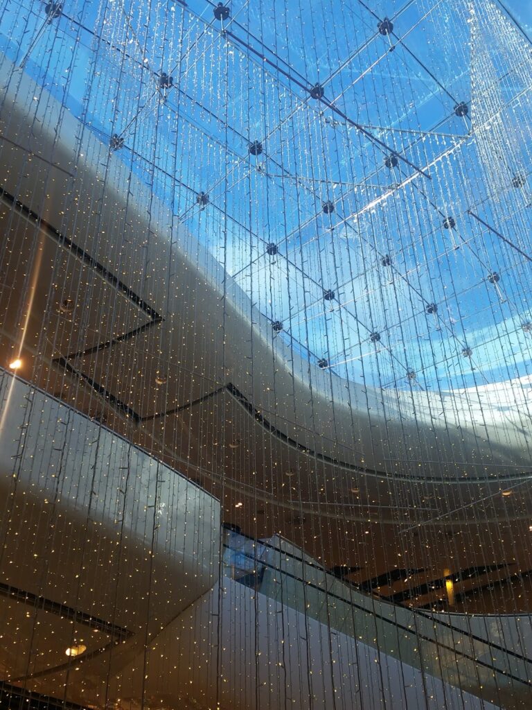 A photo taken from inside a modern building shows a high glass ceiling with a wavy design, through which blue sky and clouds are visible. Strings of small, warm white lights hang from the ceiling, creating a sparkling, elegant effect.