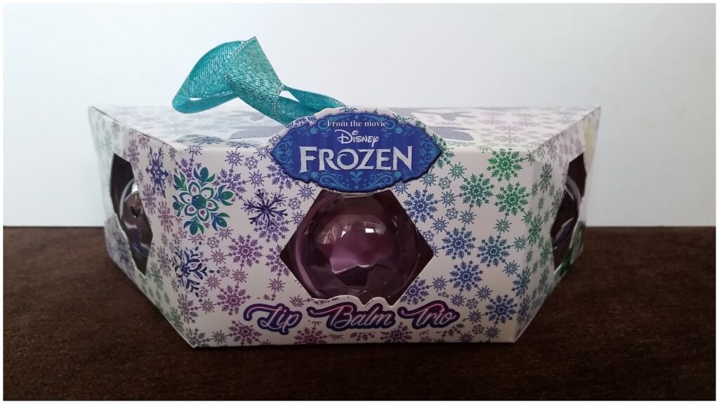 The image shows a Frozen-themed lip balm trio set in snowflake-patterned packaging. There's a blue-green ribbon on top and a purple lip balm visible through a window. The packaging features the Disney Frozen logo and "Lip Balm Trio" text in purple.