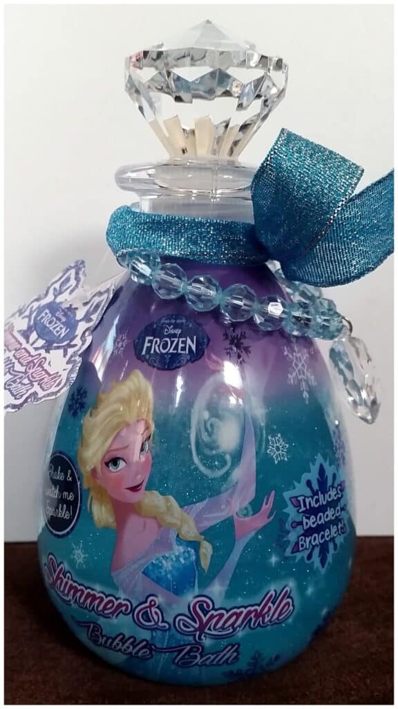 A bottle of Disney Frozen Shimmer & Sparkle Bubble Bath shaped like a potion vial, with a faceted plastic crystal cap, blue ribbon, and bead bracelet. The label features Elsa, snowflakes, and purple and blue colours, promoting included bracelet inside.