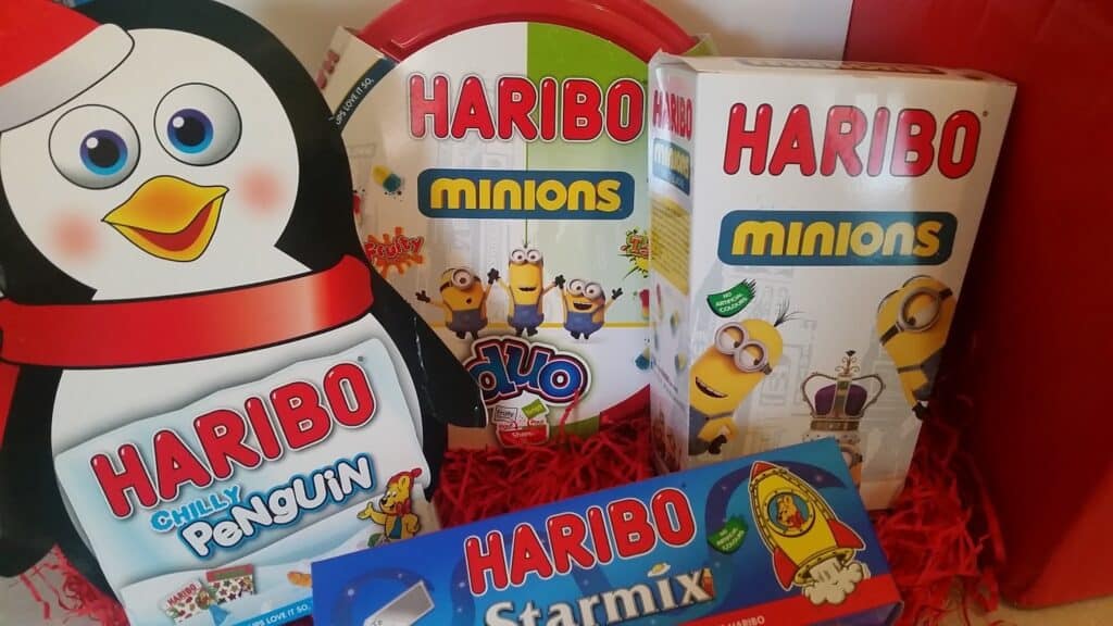 A festive display of Haribo sweet boxes, including a penguin-shaped "Chilly Penguin" box, two "Minions" boxes with Minion characters, and a "Starmix" box with a rocket image, all set on red shredded paper.