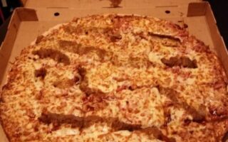 An open pizza box shows a large cheese pizza topped with uneven strips of breaded chicken. There is a large grease stain in the shape of a heart on the inside of the box lid above the pizza.