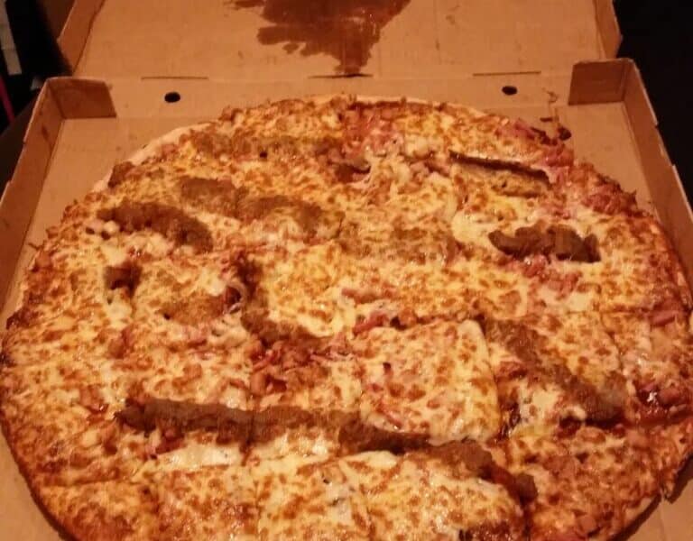An open pizza box shows a large cheese pizza topped with uneven strips of breaded chicken. There is a large grease stain in the shape of a heart on the inside of the box lid above the pizza.